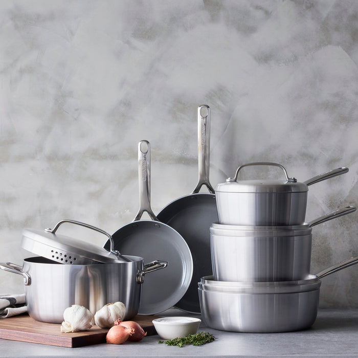 Stainless steel 10-piece cookware set with mirror handles on a counter, surrounded by garlic, shallots, herbs, and a bowl.