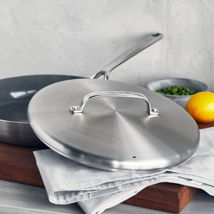 Stainless steel frypan with lid and mirror handles on a cloth, next to a lemon, herbs, and a small bowl.