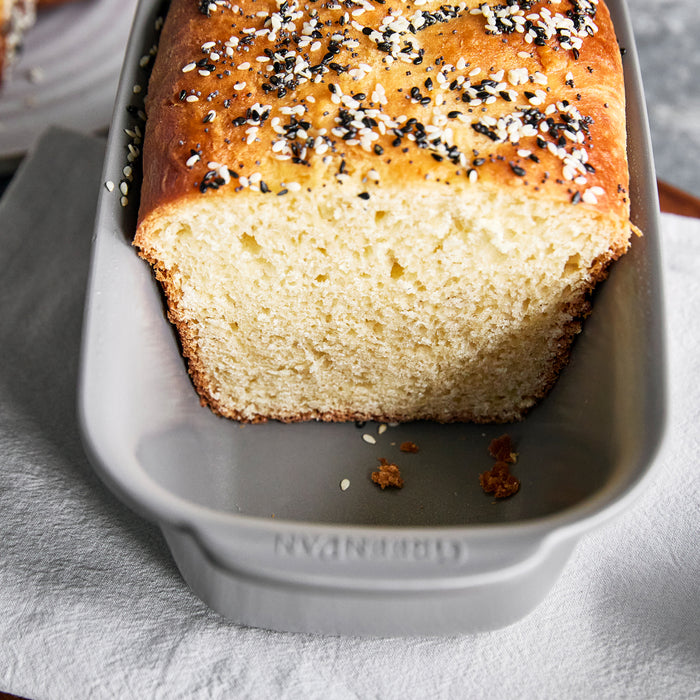 Gray ceramic 1 lb. loaf pan holding a partially sliced seeded bread loaf, placed on a white cloth.