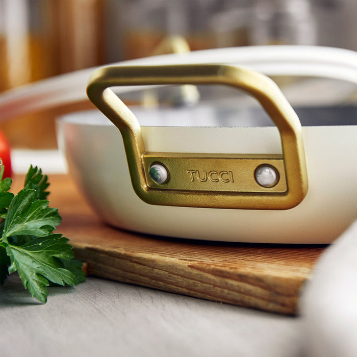 White ceramic 12-inch frypan with gold handle, Stanley Tucci brand, placed on a board near fresh herbs.