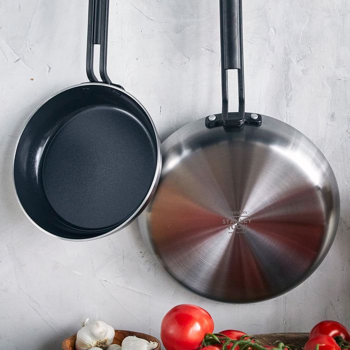 Two stainless steel nonstick frypans, one facing up and one down, with tomatoes and garlic on a neutral background.