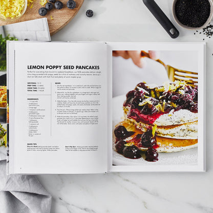 Cookbook open to a lemon poppy seed pancake recipe with pancakes topped with whipped cream, blueberries, and lemon zest.