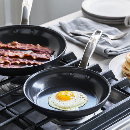 Fried egg and bacon cooking in a black ceramic 8-inch frypan on a stove, with plates next to the pan.