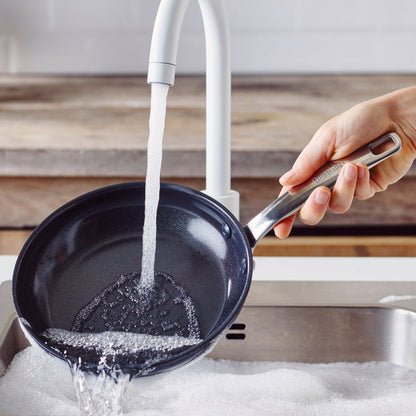 Hand rinsing a black ceramic 8-inch frypan under running water in a soapy kitchen sink.