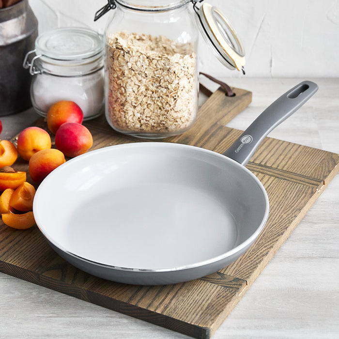 Charcoal gray ceramic nonstick frypan on a board with apricots, oats, and glass jars nearby.