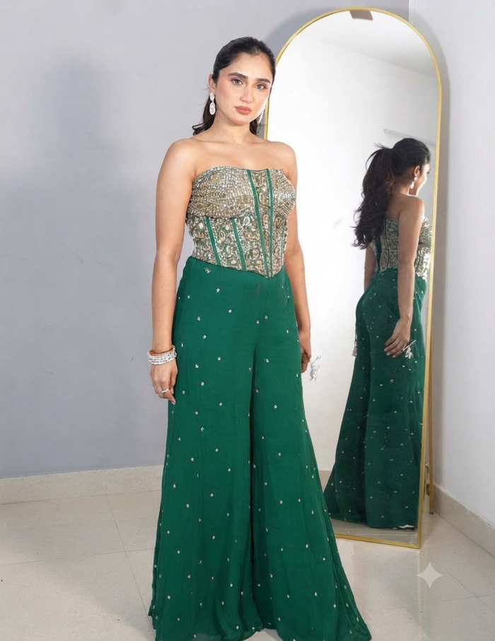 A woman with her hair tied in a ponytail wearing a strapless green embroidered top and matching wide-leg trousers, accessorized with earrings and bracelets, standing in front of a mirror.
