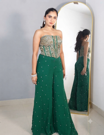 A woman with her hair tied in a ponytail wearing a strapless green embroidered top and matching wide-leg trousers, accessorized with earrings and bracelets, standing in front of a mirror.