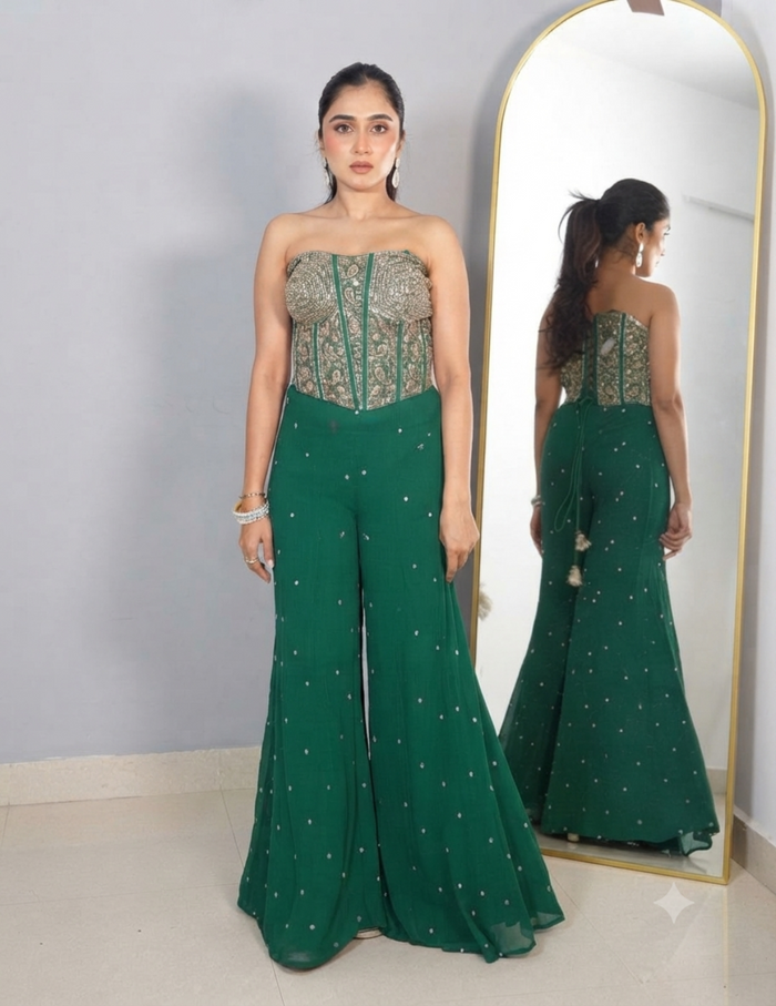 A woman with her hair tied in a ponytail wearing a strapless green embroidered top and matching wide-leg trousers, accessorized with earrings and bracelets, standing in front of a mirror.