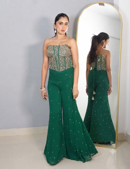 A woman with her hair tied in a ponytail wearing a strapless green embroidered top and matching wide-leg trousers, accessorized with earrings and bracelets, standing in front of a mirror.