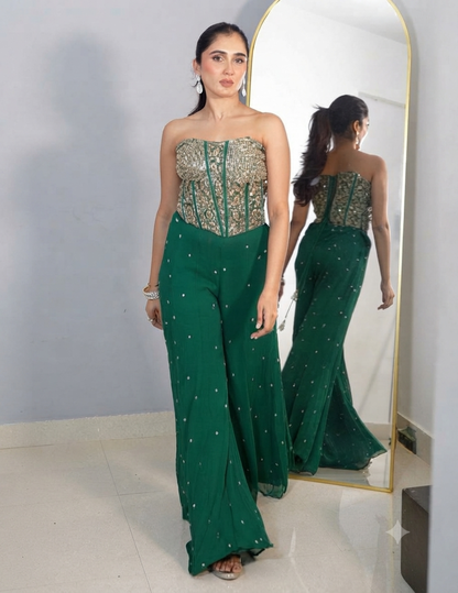 A woman with her hair tied in a ponytail wearing a strapless green embroidered top and matching wide-leg trousers, accessorized with earrings and bracelets, standing in front of a mirror.