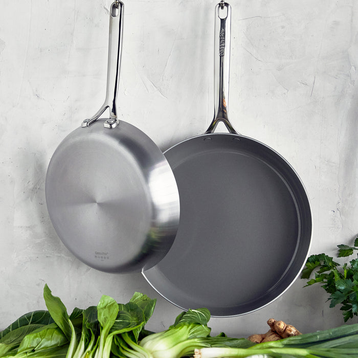 Two stainless steel frypans with mirror handles hang above fresh greens and ginger.