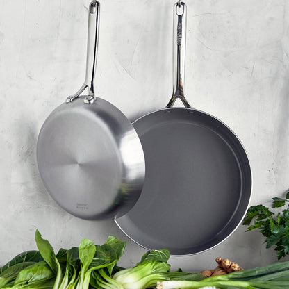 Two stainless steel frypans with mirror handles hang above fresh greens and ginger.