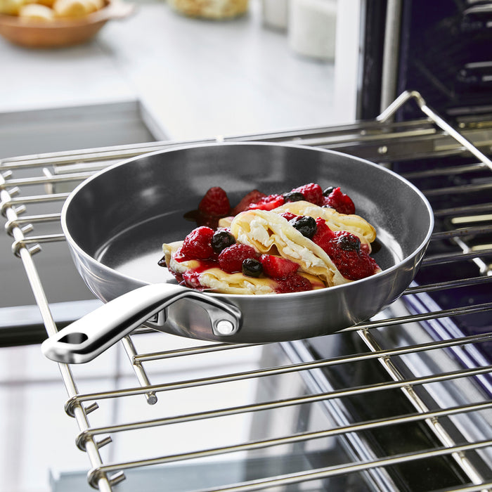 Set of two stainless steel frypans with mirror handles, each holding pancakes and berries, on an oven rack.