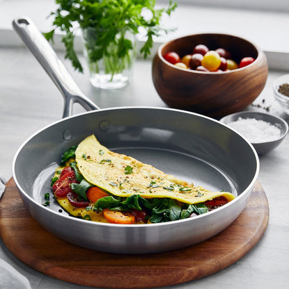 Stainless steel frypan with an omelette, spinach, and tomatoes; herbs and bowls blurred in the background.
