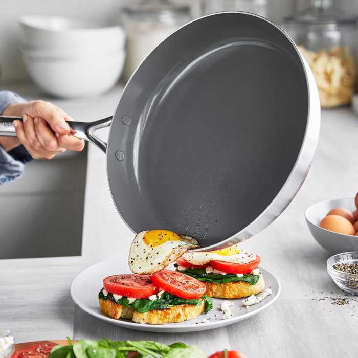 A stainless steel frypan slides a fried egg onto toast topped with spinach, tomato, and cheese.