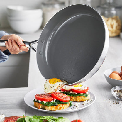 A stainless steel frypan slides a fried egg onto toast topped with spinach, tomato, and cheese.