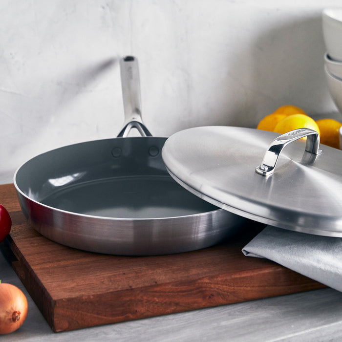 Stainless steel 12-inch frypan with lid and mirrored handles on a wooden board, lemons and bowls in background.