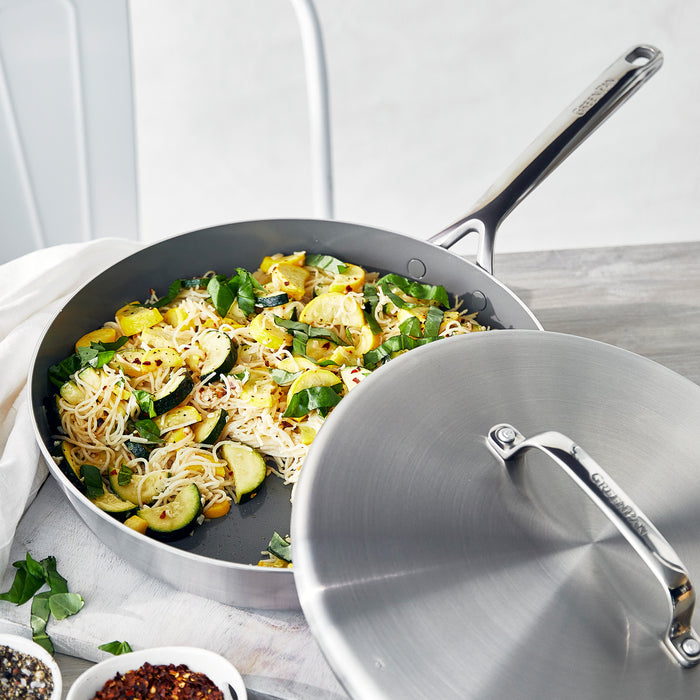 Stainless steel 12-inch frypan with lid and mirror handles, filled with noodles, zucchini, and spinach on a light surface.