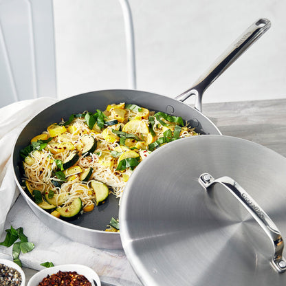 Stainless steel 12-inch frypan with lid and mirror handles, filled with noodles, zucchini, and spinach on a light surface.