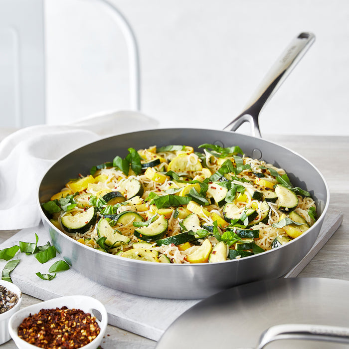 Stainless steel frypan with lid containing pasta, zucchini, and herbs on a light wood table.