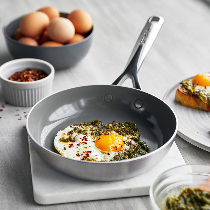 Stainless steel 8-inch frypan with mirror handle, cooking a fried egg with herbs, next to eggs and toast.