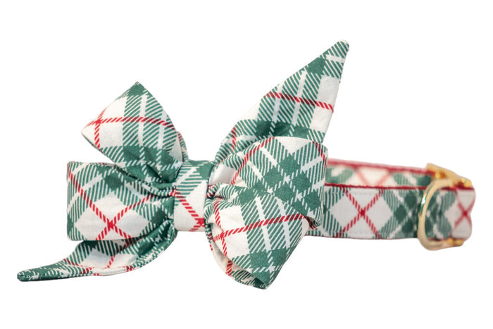 A festive dog collar with an attached oversized matching belle bow in green plaid on a light fabric background. The belle bow features structured loops and pointed tails, and the collar includes a  metal buckle and D-ring, combining playful holiday charm with elegant details.