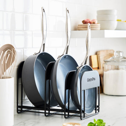 Three frying pans in a GreenPan rack on a kitchen counter, with utensils and jars visible in the background.