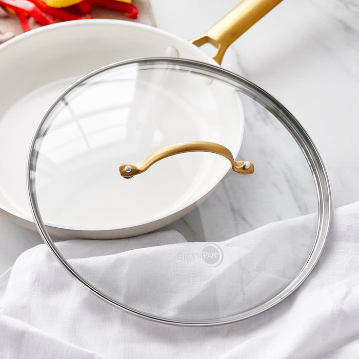 Tempered glass lid with gold knob next to a white frying pan with gold handle on a white surface.