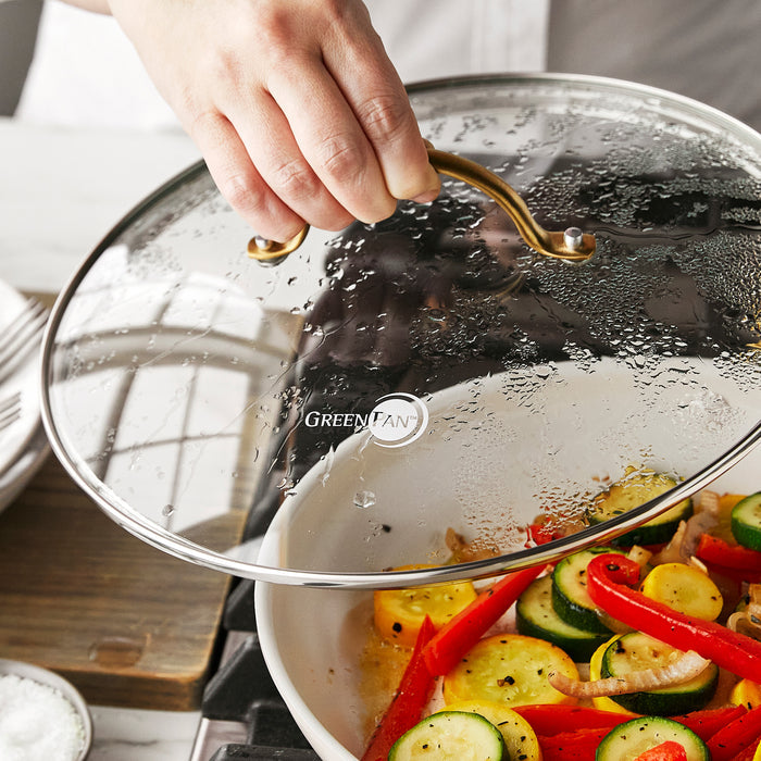 Hand lifting a GreenPan glass lid with matte gold handle from a pan of sliced vegetables.