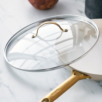 Glass lid with matte gold handle resting on a white pan, set on a marble countertop.