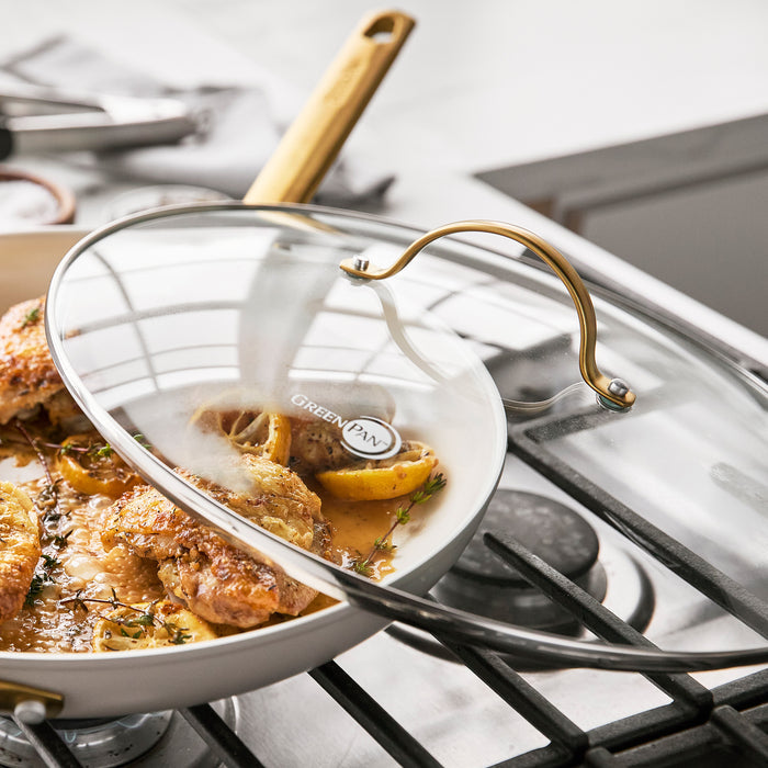Clear glass lid with stainless steel handle covering a skillet with chicken on a gas stove in a bright kitchen.