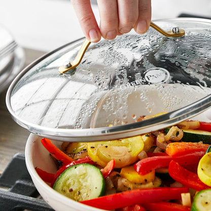 A hand lifts an 8-inch clear glass lid with black handle from a pan of sautéed zucchini and bell peppers.