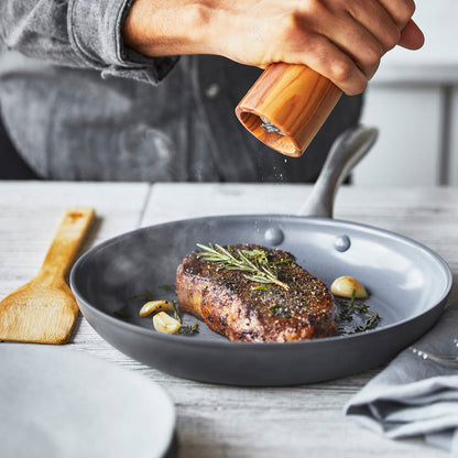 Pepper being ground over steak in a green ceramic frying pan with bamboo spatula on a light wood table.