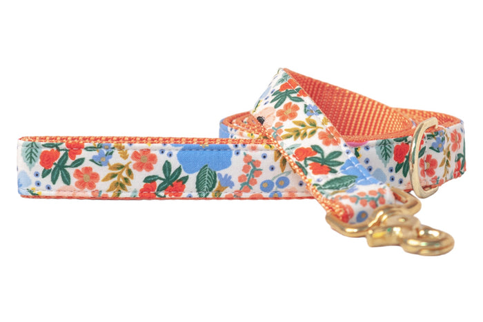 A fabric dog leash with a colorful floral print featuring red, orange, blue, and green flowers on a white background. It has a peachy orange webbing lining, a gold metal clasp, and a matching D-ring.