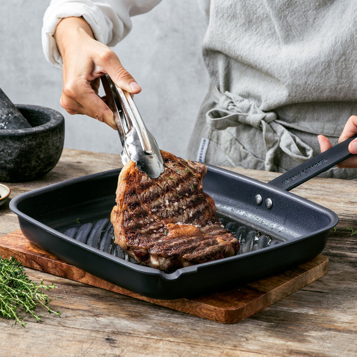 Grilled steak lifted with tongs from a black carbon steel square grill pan on a table.