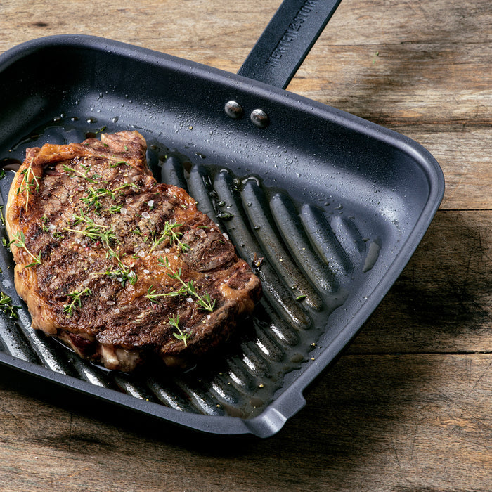 Grilled steak with herbs in a black carbon steel square grill pan on a rustic wooden table.
