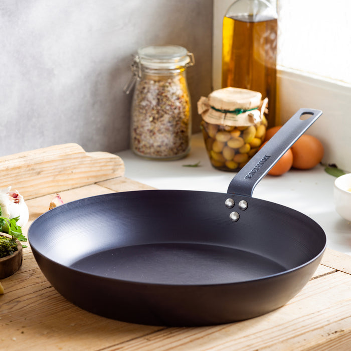 Carbon steel 12-inch frypan on a wooden counter next to glass jars filled with grains, olives, and olive oil.