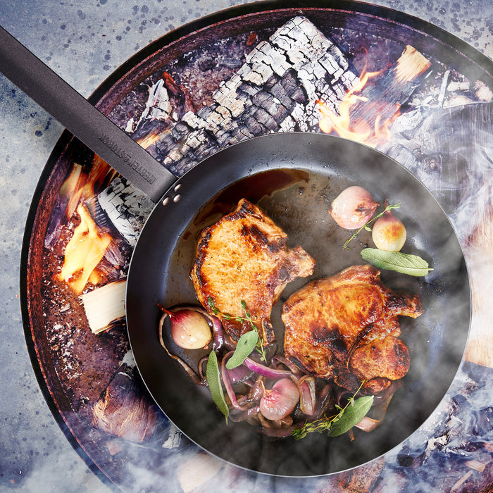 Carbon steel frypan cooking pork chops, onions, and herbs over an open flame with smoke rising.