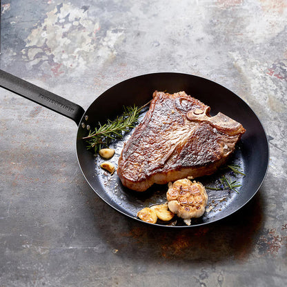 T-bone steak with garlic and rosemary cooking in a Merten  Storck carbon steel frypan on a rustic surface.