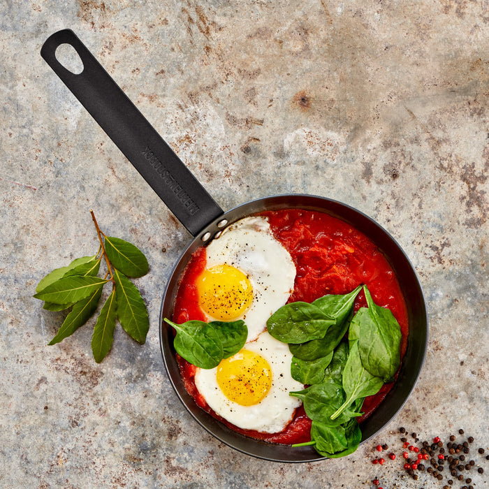 Two black carbon steel frying pans with tomato sauce, fried eggs, spinach, and bay leaves on a stone surface.