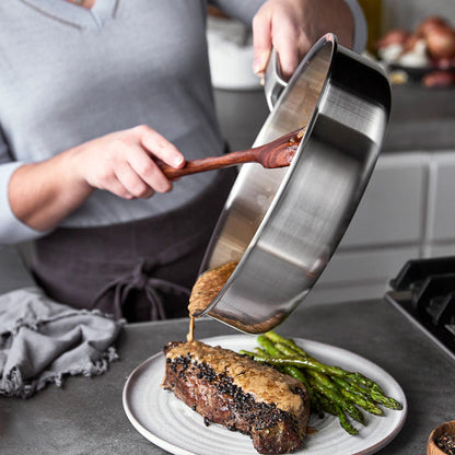 Sauce being poured in a blue Merten  Storck sauté pan onto steak and asparagus.