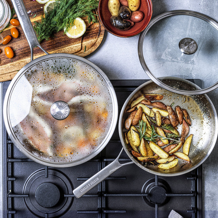 Stainless steel sauté pan with lid on stovetop, potatoes and herbs cooking nearby.