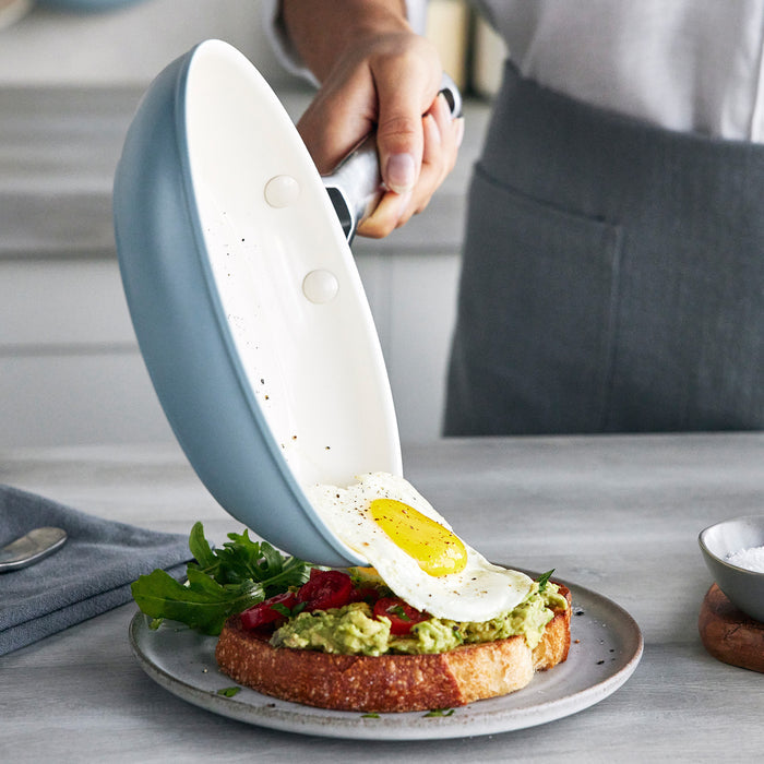 A fried egg being slid from a gray ceramic nonstick frypan onto avocado toast with greens and tomatoes.