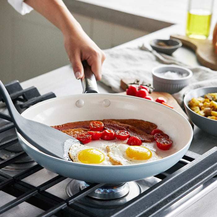 Two green ceramic nonstick frypans with eggs, bacon, and tomatoes cooking on a stove, spatula in use. Set of 2.