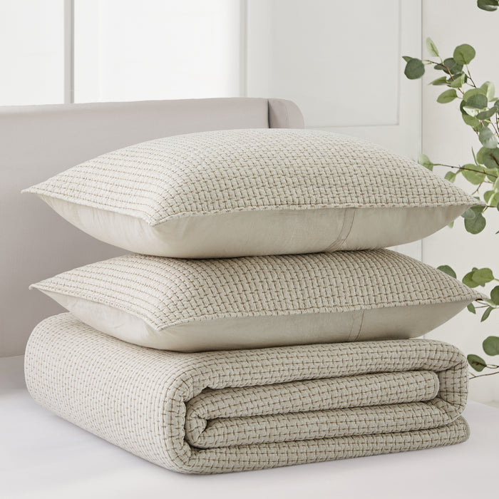 Folded beige textured blanket with two pillows on a light-colored surface, with a blurred plant in the background.