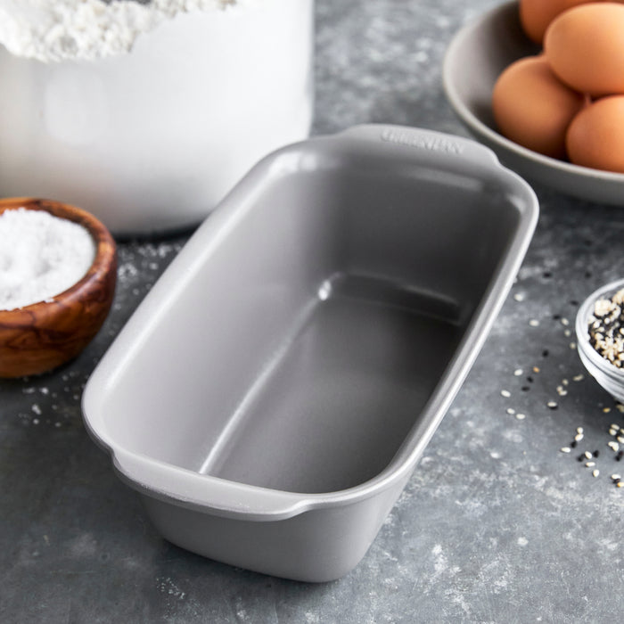 Gray ceramic nonstick loaf pan on a countertop with eggs, flour, salt, and seeds nearby.