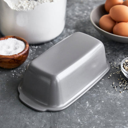 Gray ceramic loaf pan, upside down on a counter with eggs, flour, salt, and seeds nearby.