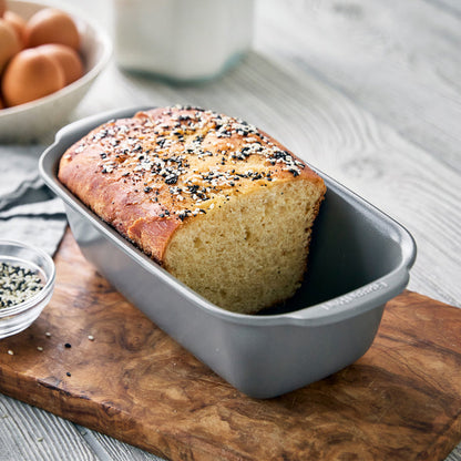 Gray ceramic nonstick loaf pan with seeded bread on a wooden board, eggs and a glass of milk beside it.