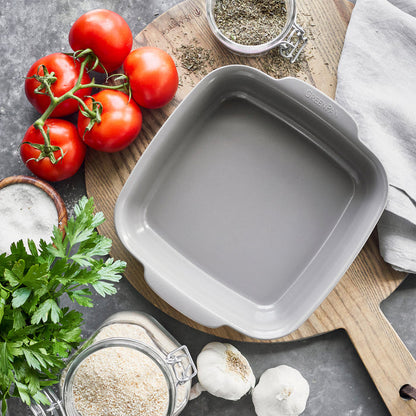 Gray ceramic nonstick square baker on a wooden board, surrounded by tomatoes, parsley, garlic, and herbs.