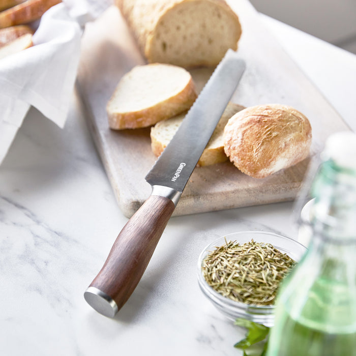 Titanium bread knife with walnut handle next to sliced bread, fresh herbs, and a green glass bottle on a table.